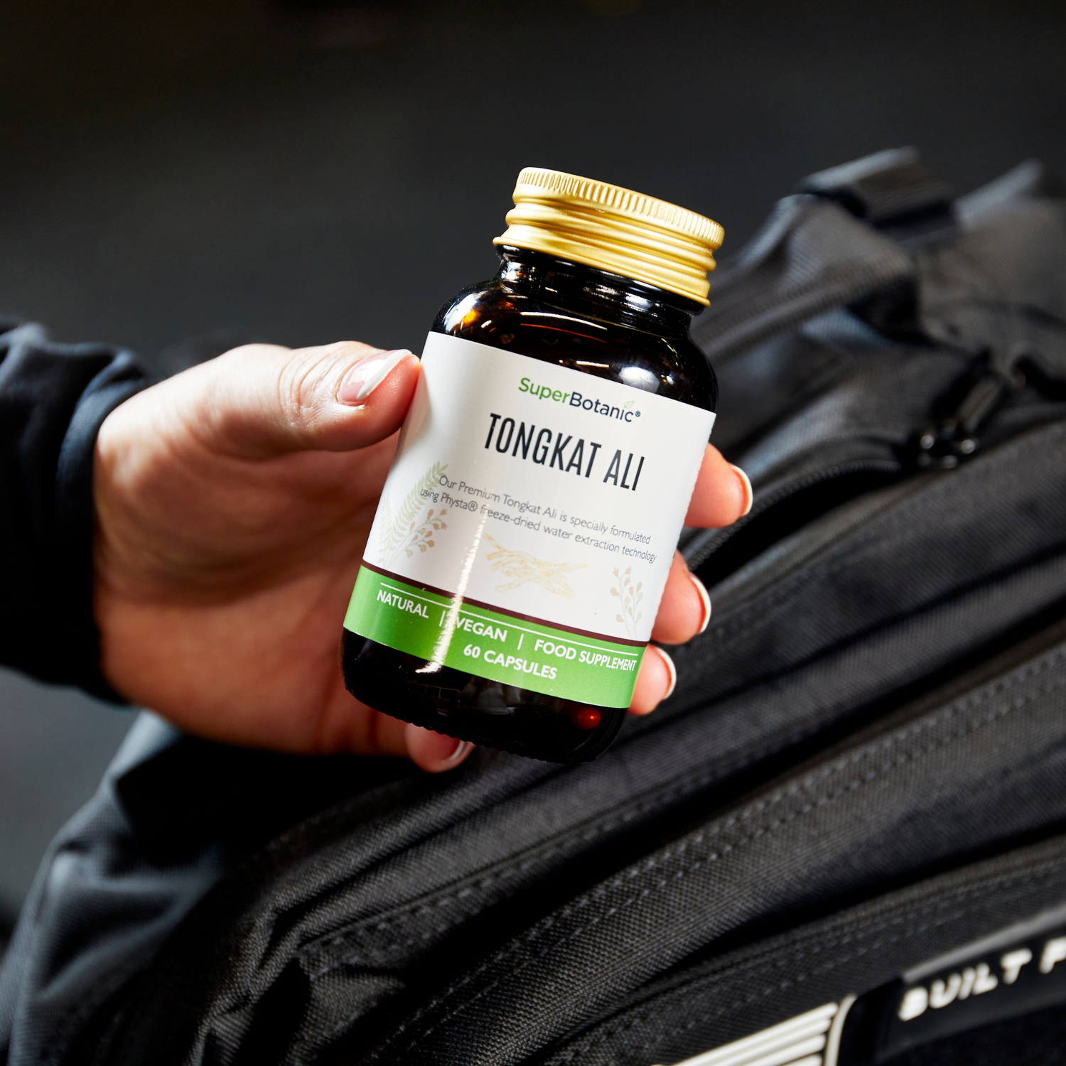 Hand holding a bottle of Tongkat Ali supplements near a black bag.